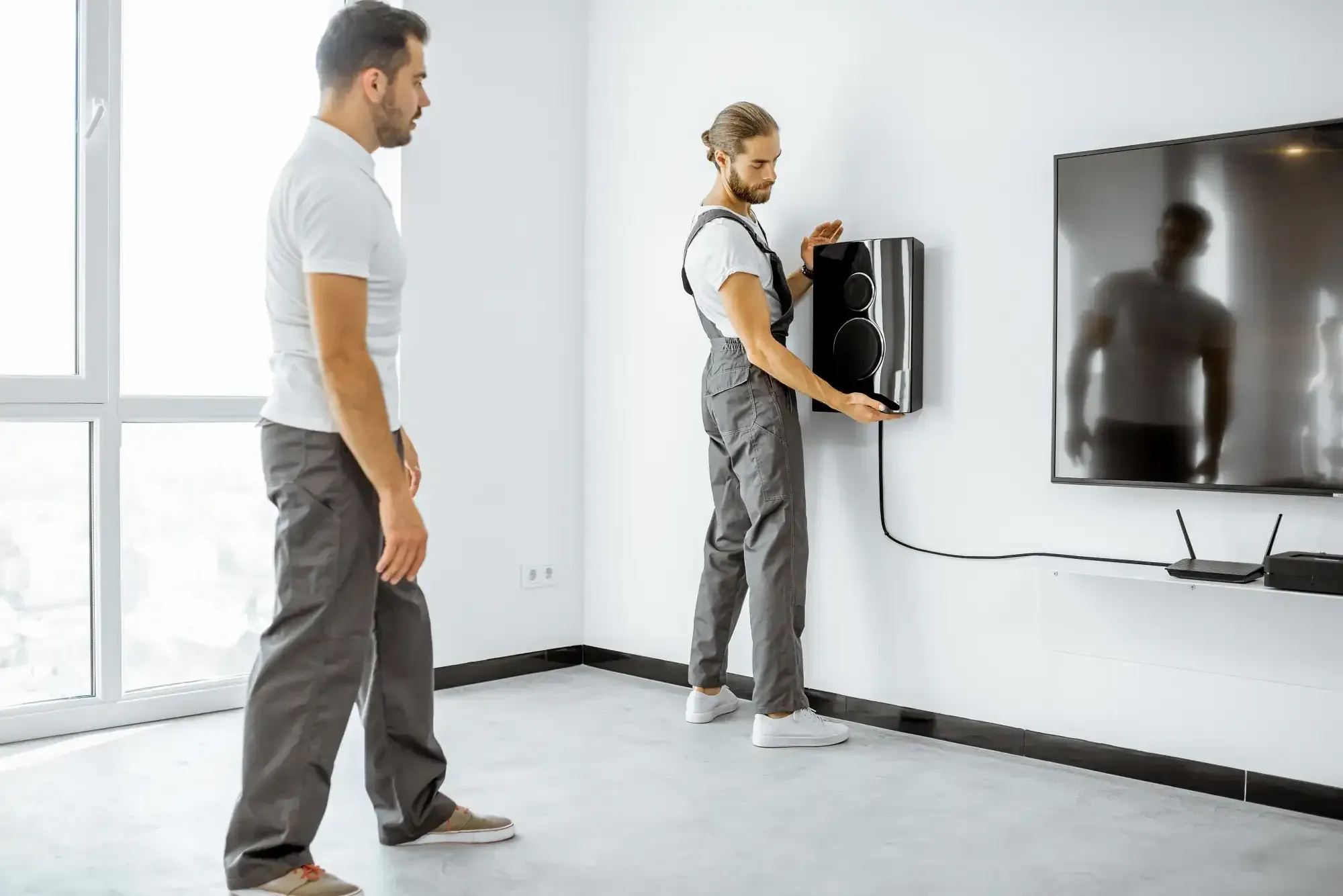 Two men installing a speaker on a white wall near a TV. One man is holding the speaker.