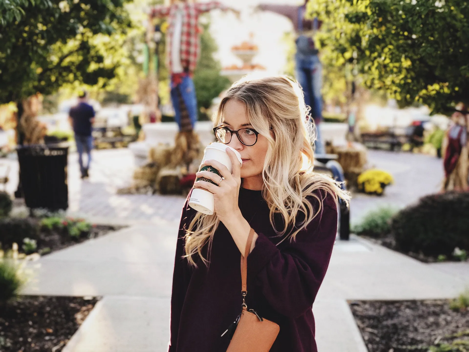 Blonde woman sipping coffee outdoors, wearing glasses and a maroon sweater in a garden setting.